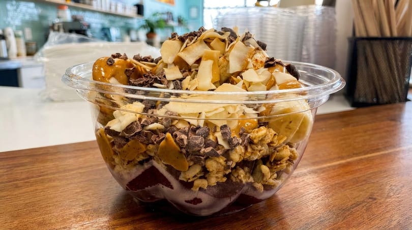 A vibrant acai bowl with granola, bananas, coconut flakes, chocolate, and peanut butter drizzle.