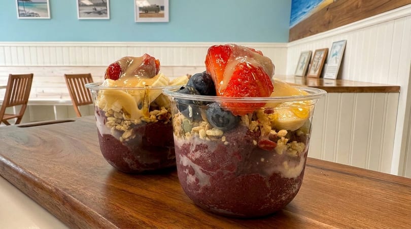 Two acai bowls topped with strawberries, blueberries, bananas, granola, and peanut butter drizzle.