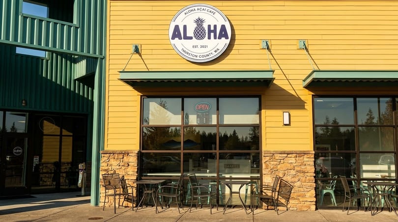 Exterior of Aloha Acai Cafe, a yellow building with a round logo, large windows, and outdoor seating.