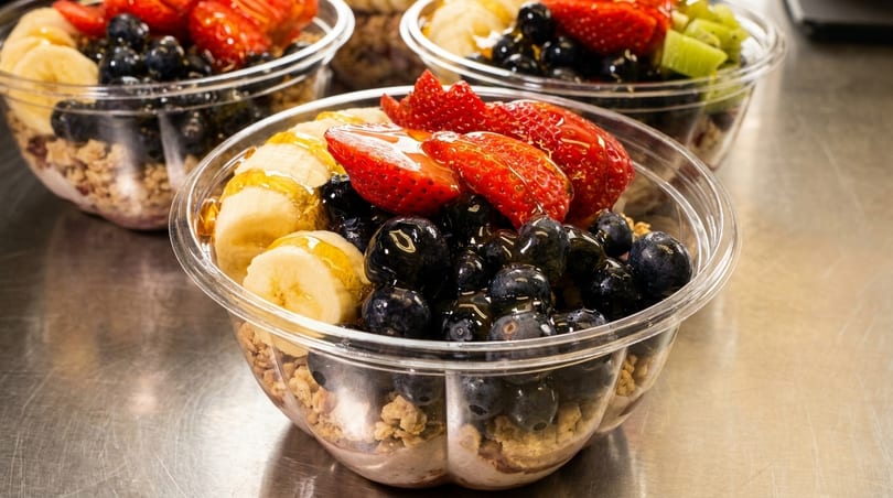 Three clear plastic bowls filled with granola, yogurt, blueberries, strawberries, bananas, and honey.