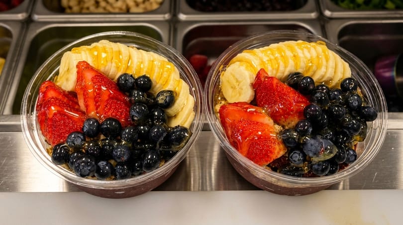 Two acai bowls topped with sliced bananas, strawberries, blueberries, and a honey drizzle.